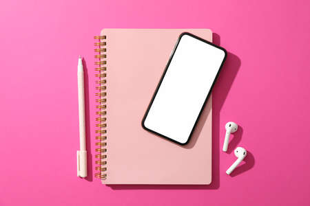 Phone with empty screen, headphones, copybook and pen on pink background, top view