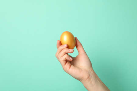 Female hand holds golden egg on mint background, space for textの写真素材