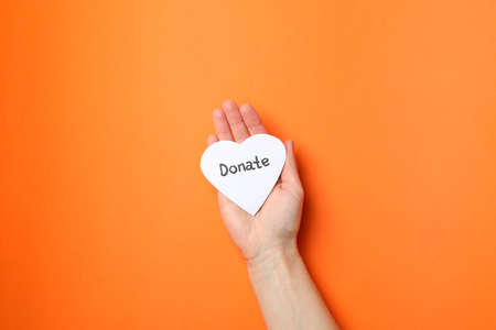 Female hand holds heart with inscription Donate on orange background, top view