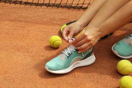 Woman tying shoelaces on clay court with racket and tennis ballsの写真素材