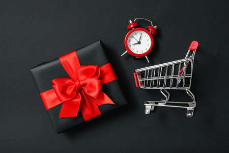 Gift box, alarm clock and shop trolley on black background