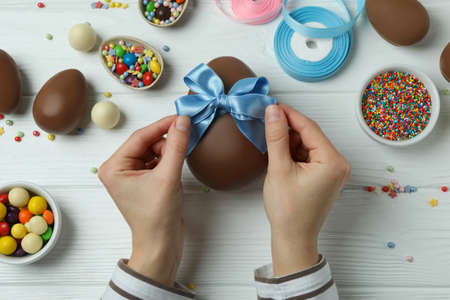 Female hands hold bow on chocolate egg on white wooden background with Easter accessories