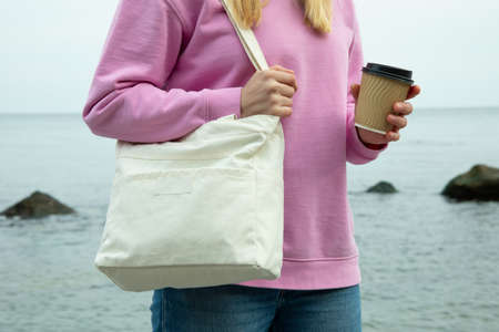Young woman hold stylish eco bag and paper cup against sea