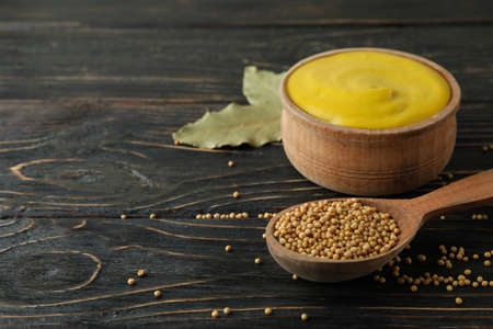 Bowl with mustard and spoon with seeds on wooden background