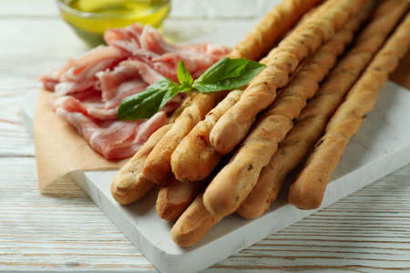 Board with grissini and bacon on white wooden background