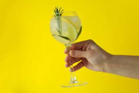Female hand hold glass of cocktail with citrus and rosemary on yellow backgroundの写真素材