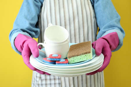 Woman in apron holds clean dishware on yellow background
