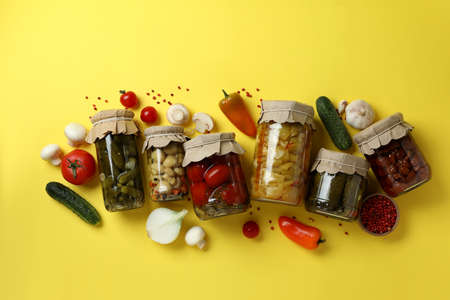 Jars of pickled vegetables and ingredients on yellow background