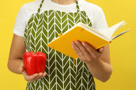 Woman holds recipe book and bell pepper on yellow backgroundの写真素材