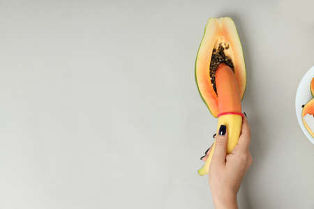 Female hand holding banana with condom in papaya on light gray backgroundの写真素材