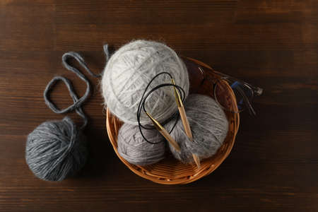 Wicker bowl with yarn balls and knitting needles, and glasses on wooden background