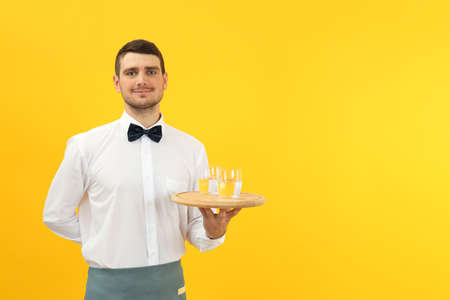 Young man waiter holds tray with glasses of water on yellow background.