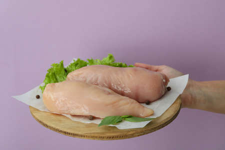 Female hand holding raw chicken fillet on wooden board on purple background