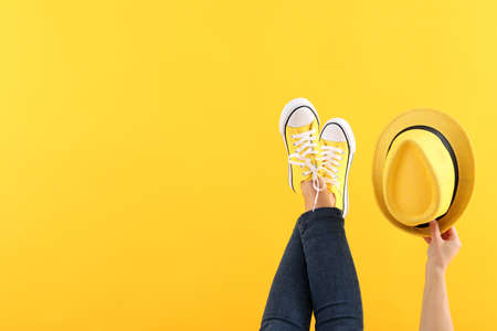 Female legs in jeans and sneakers, and female hand holding hat on yellow backgroundの写真素材