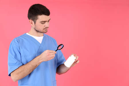 Male nurse holds bottle of pills and magnifier on pink background