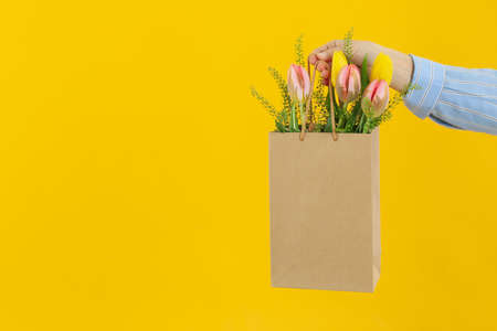 Female hand holds paper bag with flowers on yellow backgroundの写真素材