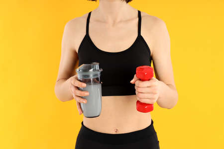 Sporty woman holds protein cocktail and dumbbell on yellow backgroundの写真素材