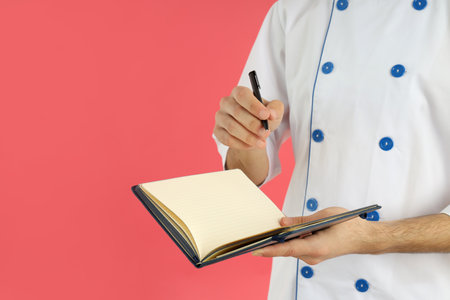 Young man chef holds recipe book and pen on pink backgroundの写真素材