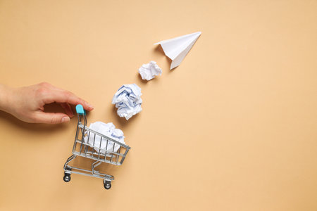 Shop cart, paper balls, paper plane and hand on beige backgroundの写真素材