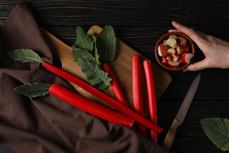 Rhubarb stalks and mint on board, brown towel, female hand, twine and knife on dark wooden...