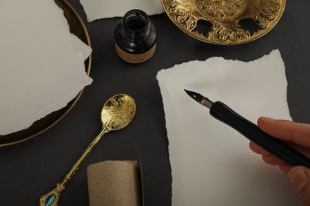 Papers, golden plates and spoon, ink bottle and pen in hand on dark gray background, top view