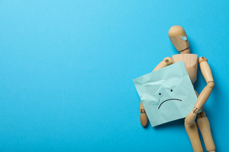 Wooden man with paper with a sad emoji on blue background, space for textの写真素材