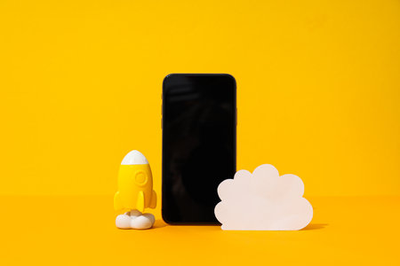 Black smartphone with paper cloud and rocket on yellow background, cloud computing concept