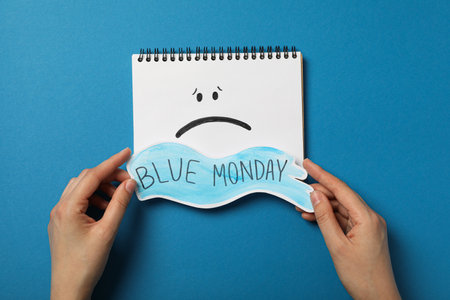 Notepad with sad emoji and text Blue Monday in hands on blue background, top viewの写真素材