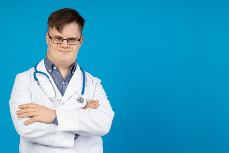 Smiling young man with cerebral palsy wearing glasses in doctor uniform with stethoscope. World Genetic Diseases Day concept, place for textの写真素材