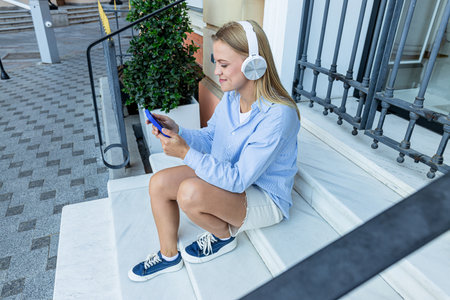 Smiling young woman in headphones and with smartphone in hands sits on steps in the cityの写真素材