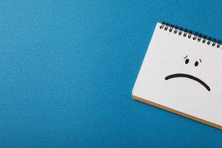 Notepad with sad emoji on blue background, space for textの写真素材