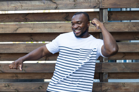 A stylish, young African-American man poses for the camera against the background of a wooden fence