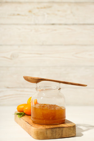 Kumquat and jar of jam on wooden stand on light wooden background, space for text