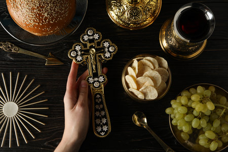 Bread, cross in hand, grapes in golden bowl and cup of wine on wooden background, top view