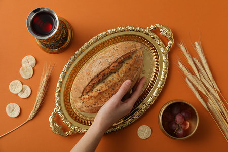 Bread on golden tray, hand, spikelets, grapes and cup of wine on orange background, top view