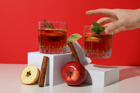Glasses with apple cider, female hand, apples and cinnamon sticks on red backgroundの写真素材