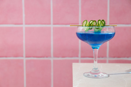 Blue cocktail with cucumber slice in glass on pink background, space for text