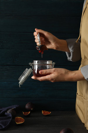 Glass jar with fig jam and spoon in female hands, towel and cut figs on table on wooden backgroundの写真素材
