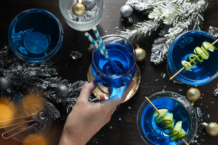 Blue cocktail in glasses, hand, christmas tree branches with snow and balls on wooden background,...