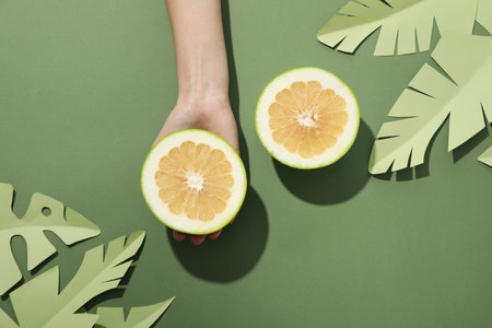 Two half of pomelo fruit, paper tropical leaves and hand on green background, top viewの写真素材