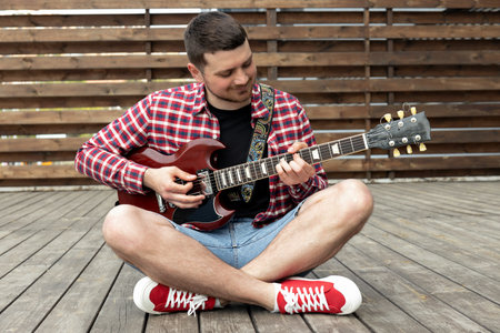 Stylish young guy playing guitar sitting on wooden stage