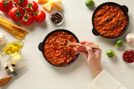 Pans with Bolognese sauce, wooden spoon in hand and ingredients on white wooden background, top viewの写真素材