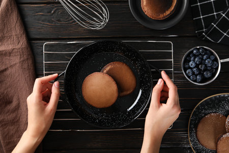 Chocolate pancakes on plates and frying pan on grid, hands, bowl with berries on wooden background,...
