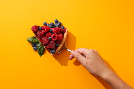 Piece of berry pie, berries, basil leaves and female hand on yellow background, top viewの写真素材