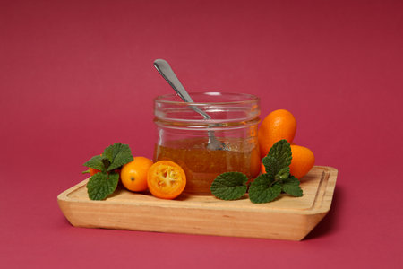 Fresh kumquat fruits, jar of jam with spoon on wooden board on pink background, close upの写真素材