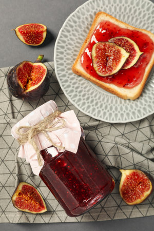 Figs, jars of jam, towel and bread with jam on plate on gray background, top view
