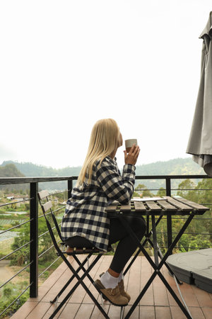 Woman sits on balcony sipping coffee with mountains in viewの写真素材