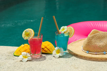 Two colorful drinks sit by a pool with fruit and flowersの写真素材