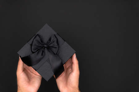 Black gift box with black satin ribbon in male hands on a black background.の写真素材