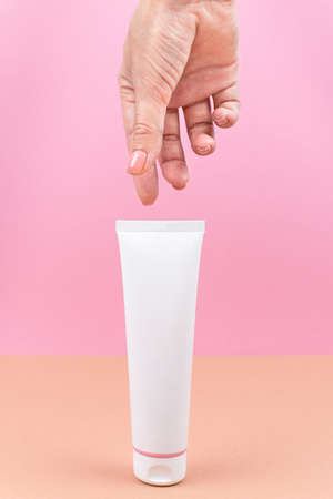 White tube with cream in a woman's hand. Closeup of cosmetic product on pink and orange background. Vertical photography. Place . Packaging of cream, lotion, gel, facial foam or skincare.の写真素材